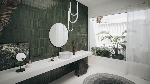 Un bagno moderno con una parete rivestita in piastrelle verde scuro, uno specchio rotondo, un piano di lavoro bianco e un'ampia finestra con piante.
