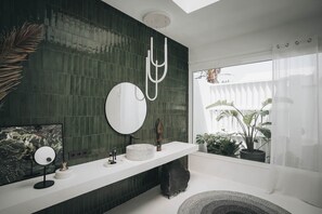 A modern bathroom with a dark green tiled wall, a round mirror, a white countertop, and a large window with plants. - Villa Amonita – Luxury Villa, Heated Pool & Private Garden (Tiagua)
