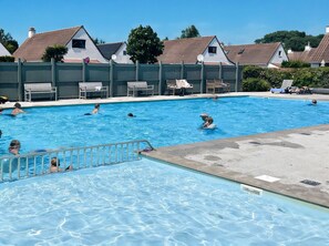 Ein Schwimmbecken mit mehreren Personen, Liegestühlen und einem Gebäude mit einem Ziegeldach im Hintergrund.
