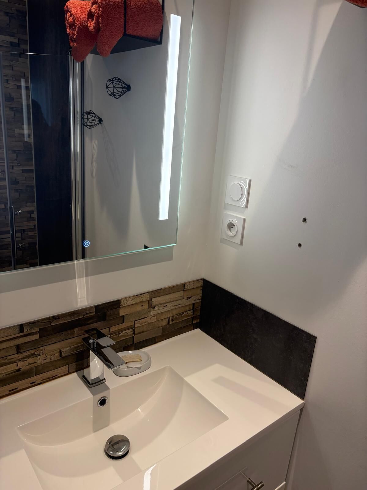Une salle de bain moderne avec une douche à l’italienne en verre, un lavabo blanc agrémenté d’un pare-éclaboussures en bois, et un miroir au-dessus du lavabo.