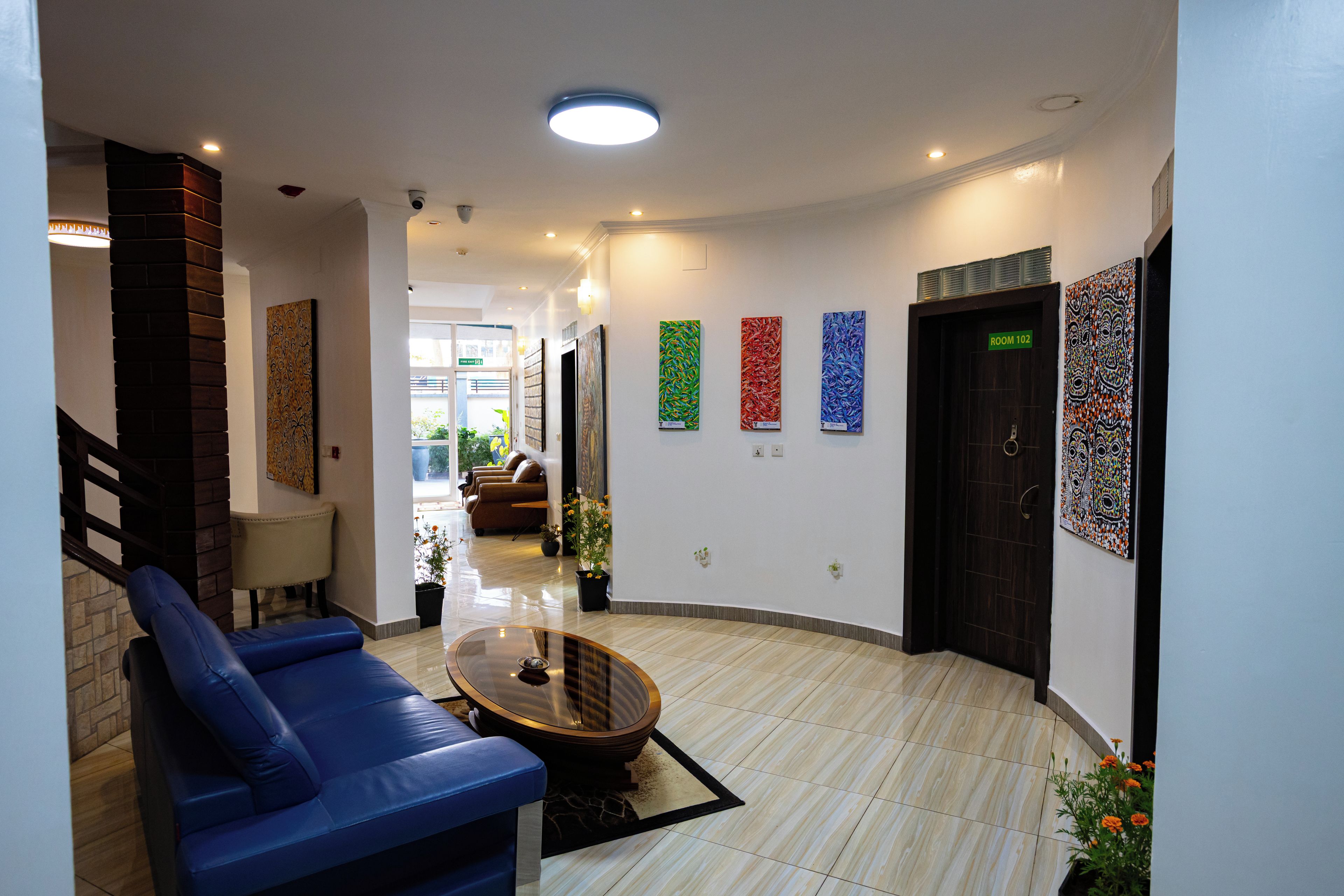 Un hall de hotel con distribución curva, una recepción, una zona de estar con sillas azules y una mesa de centro.