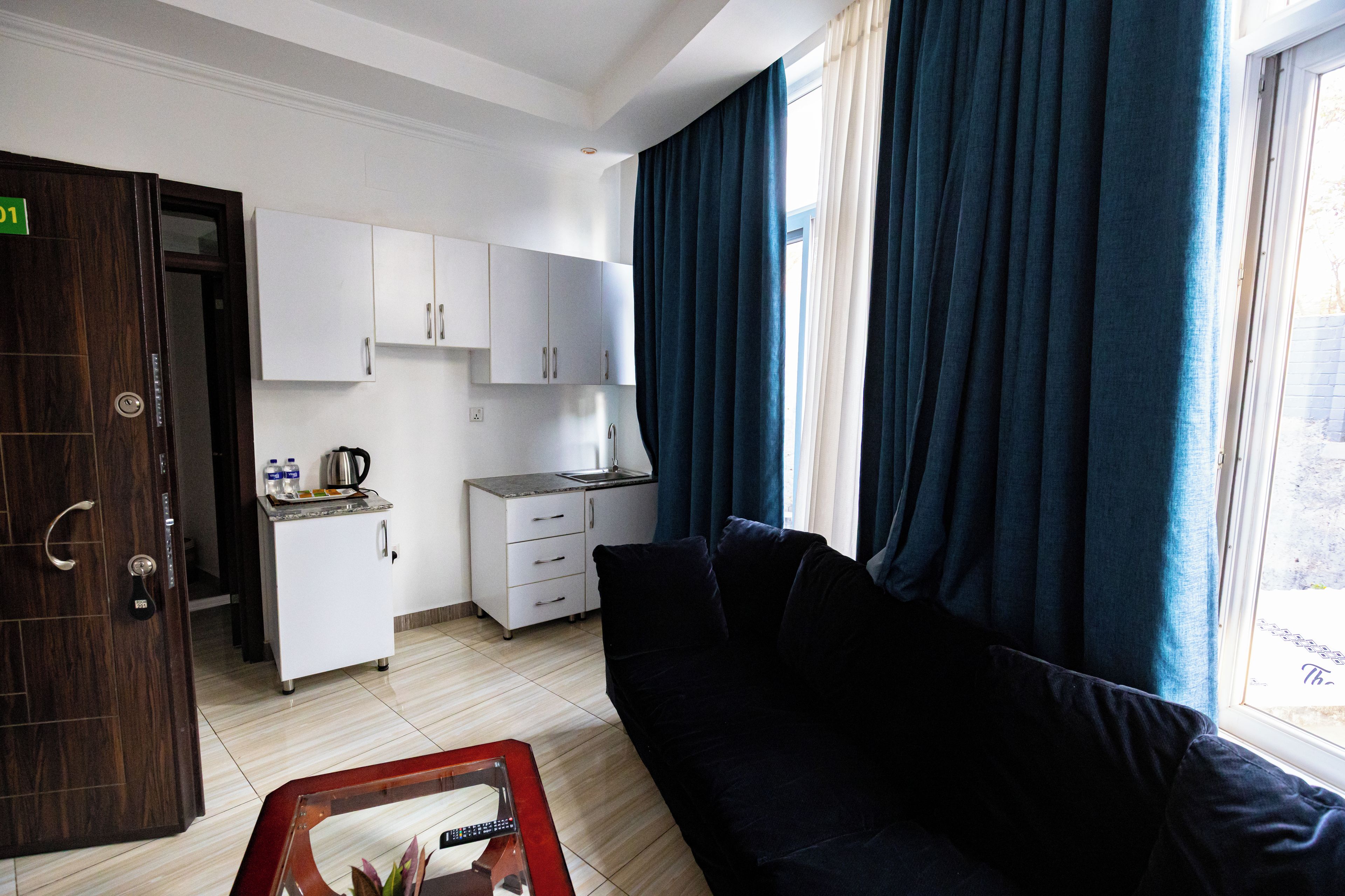 Habitación de hotel compacta con kitchenette, un sofá oscuro y un ventanal con cortinas.