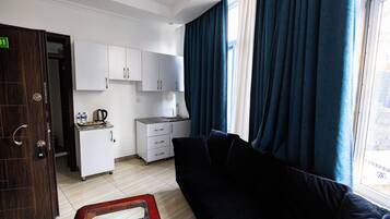 Habitación de hotel compacta con kitchenette, un sofá oscuro y un ventanal con cortinas.