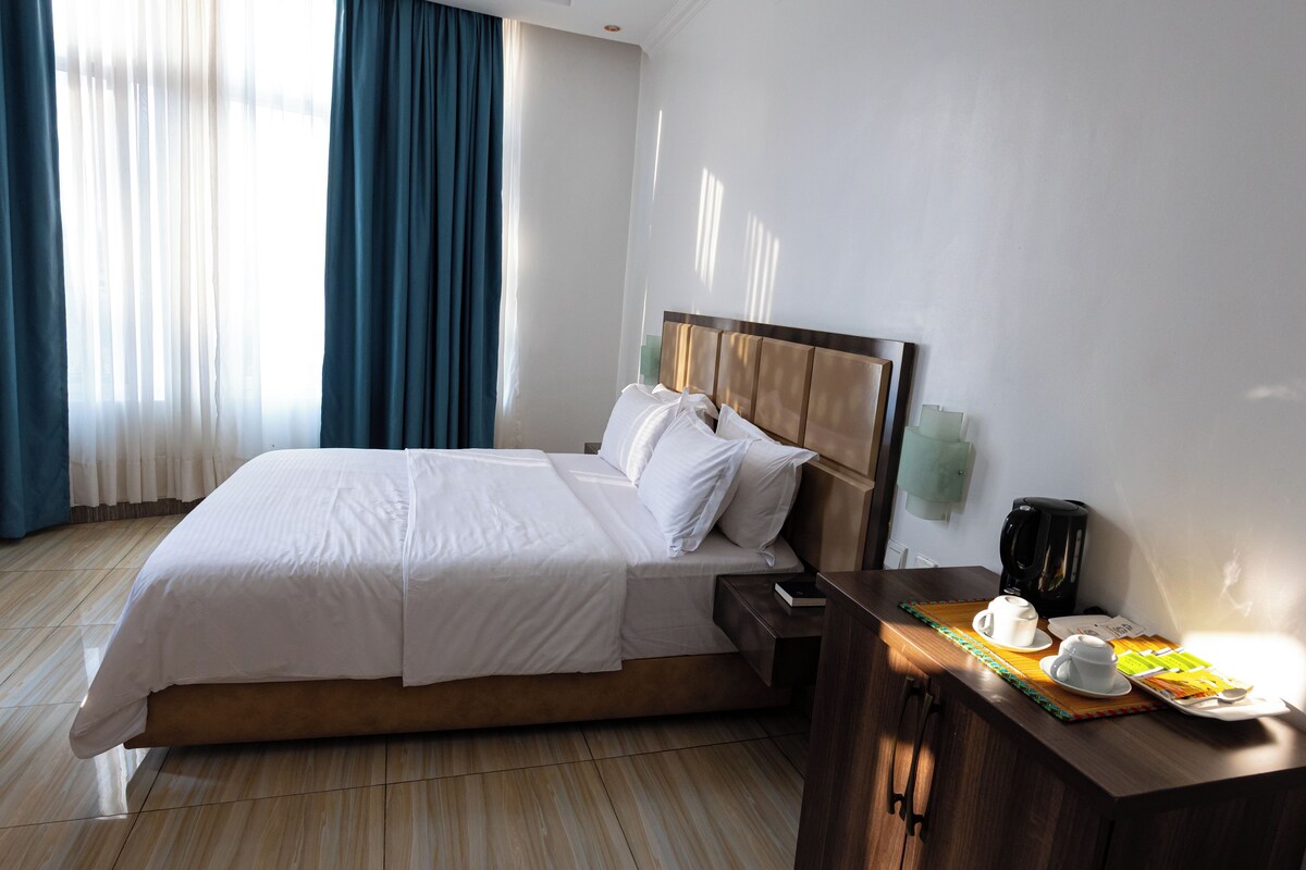 Ein Hotelzimmer mit einem Bett, einem Nachttisch mit Kaffeemaschine und einem Fenster mit Vorhängen.