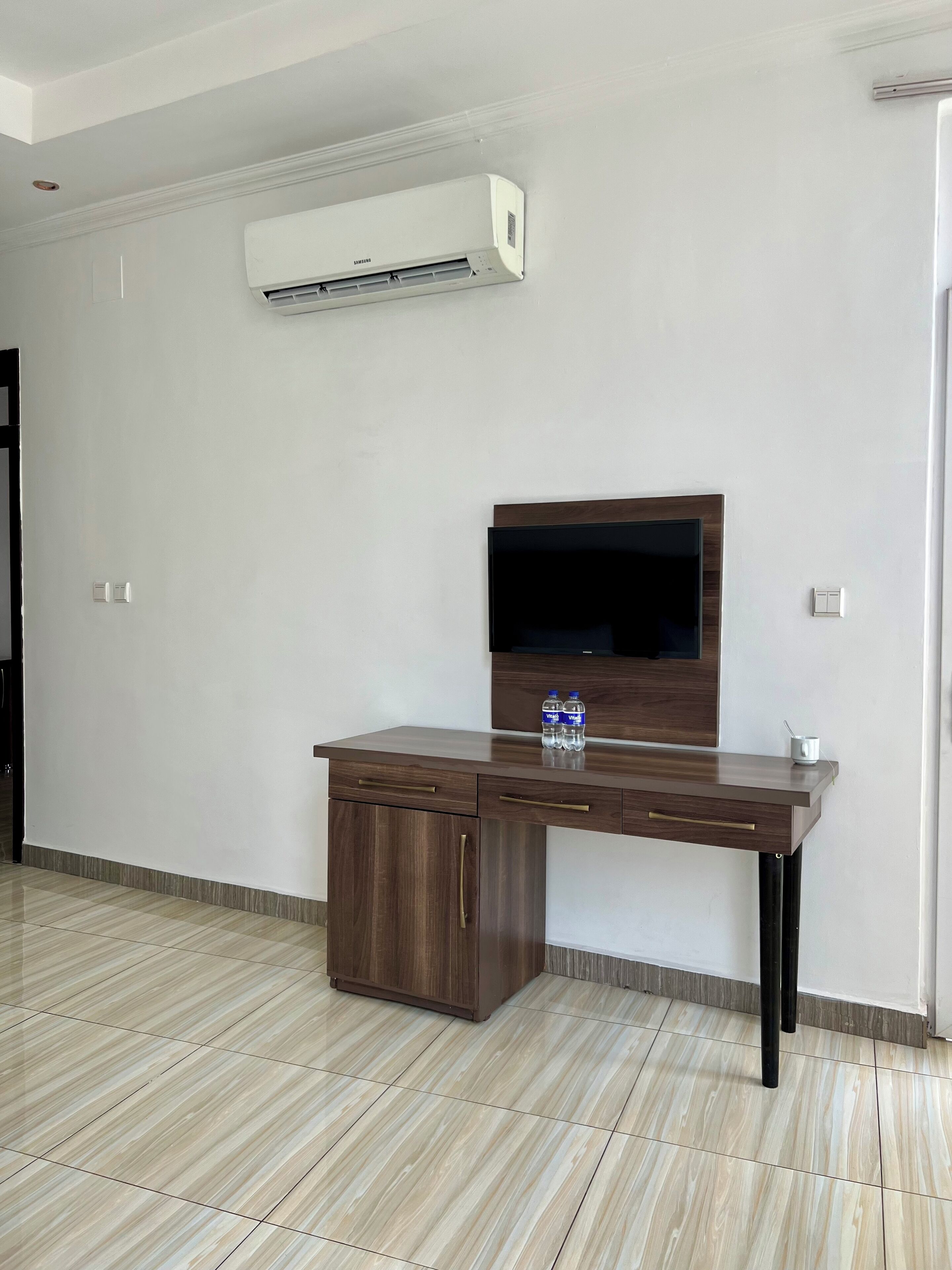 A room with a flat-screen TV mounted on a wooden cabinet, an air conditioning unit above, and a tiled floor.