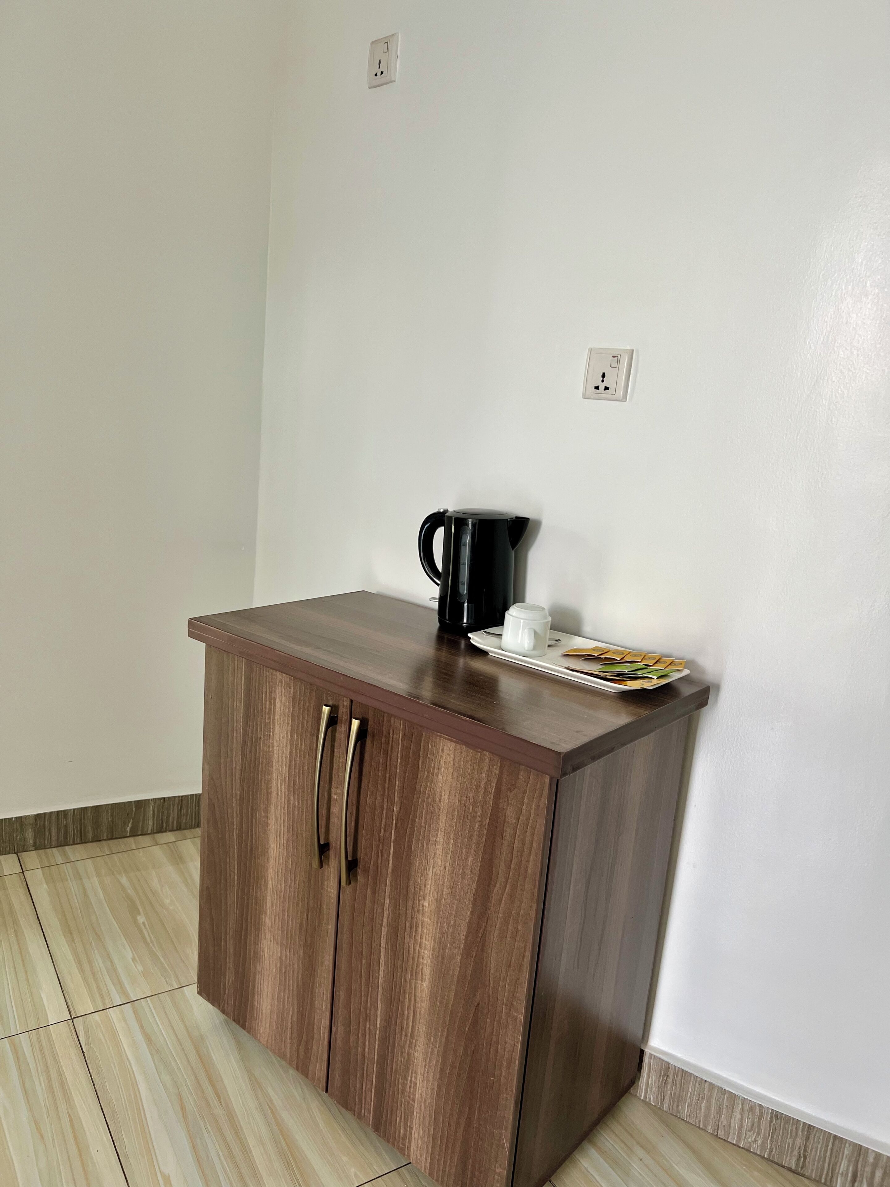 A small wooden cabinet with two drawers and a countertop, a black coffee pot, a white mug, and a plate with a sandwich on a white wall.
