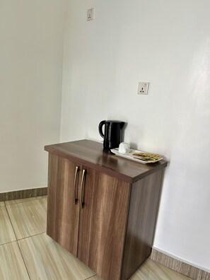 A small wooden cabinet with two drawers and a countertop, a black coffee pot, a white mug, and a plate with a sandwich on a white wall. - Eagle View Kimi (Kigali)