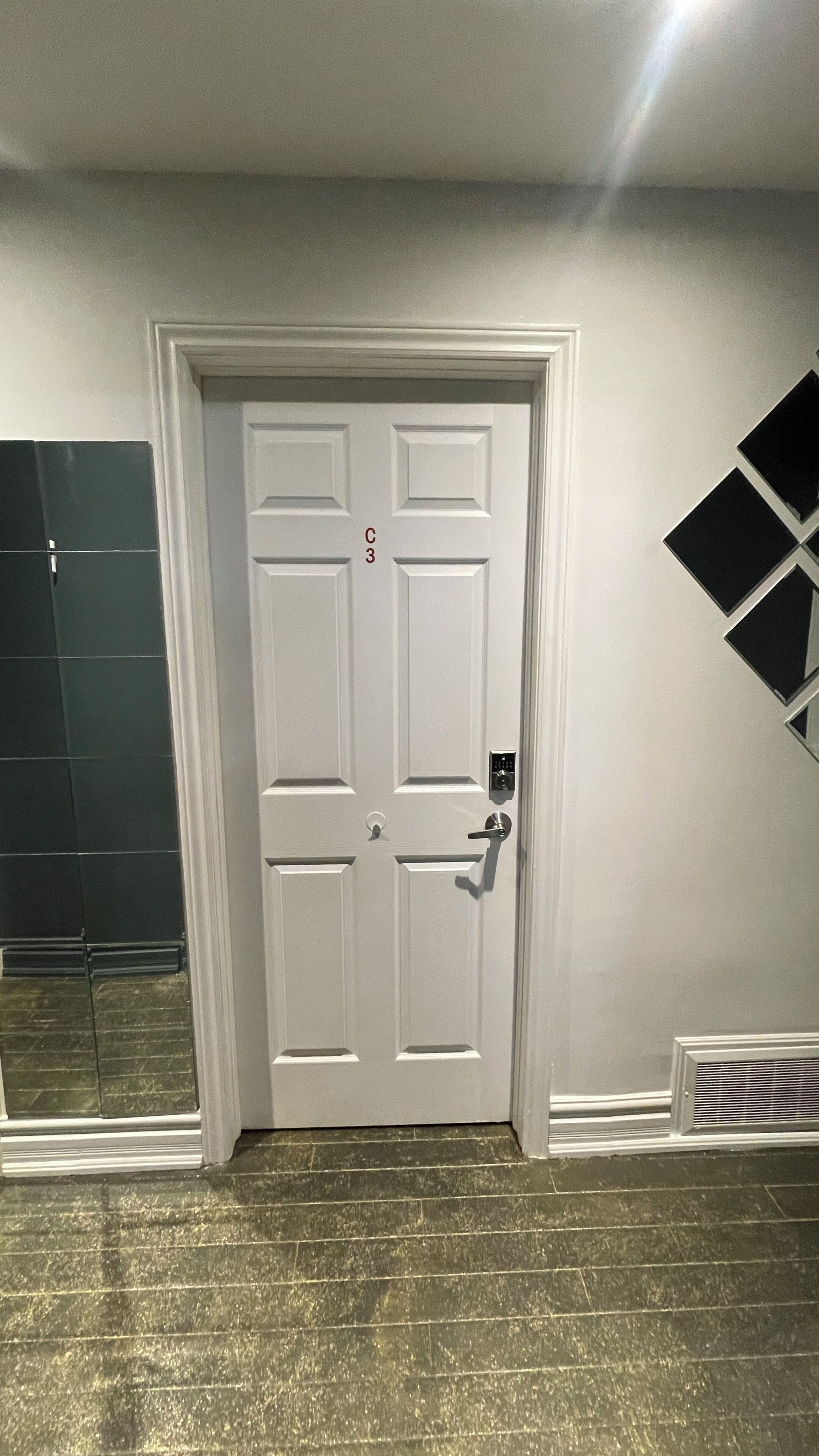 A white door with a numeric keypad lock, a vent on the floor, and a black geometric decoration on the wall.