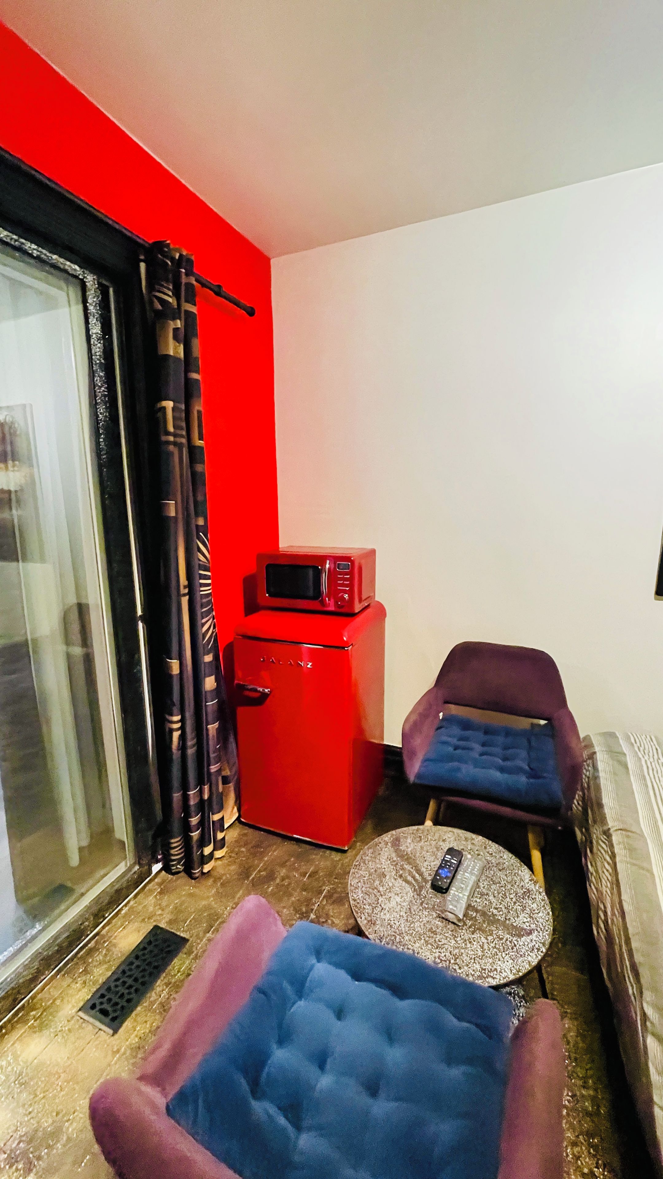 A compact living space with a red refrigerator, a microwave, a blue cushioned chair, a small round table, and a sliding glass door.