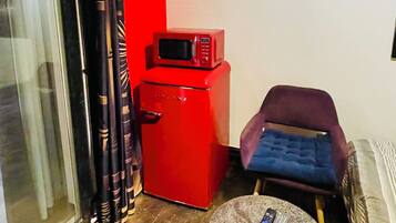 A compact living space with a red refrigerator, a microwave, a blue cushioned chair, a small round table, and a sliding glass door.