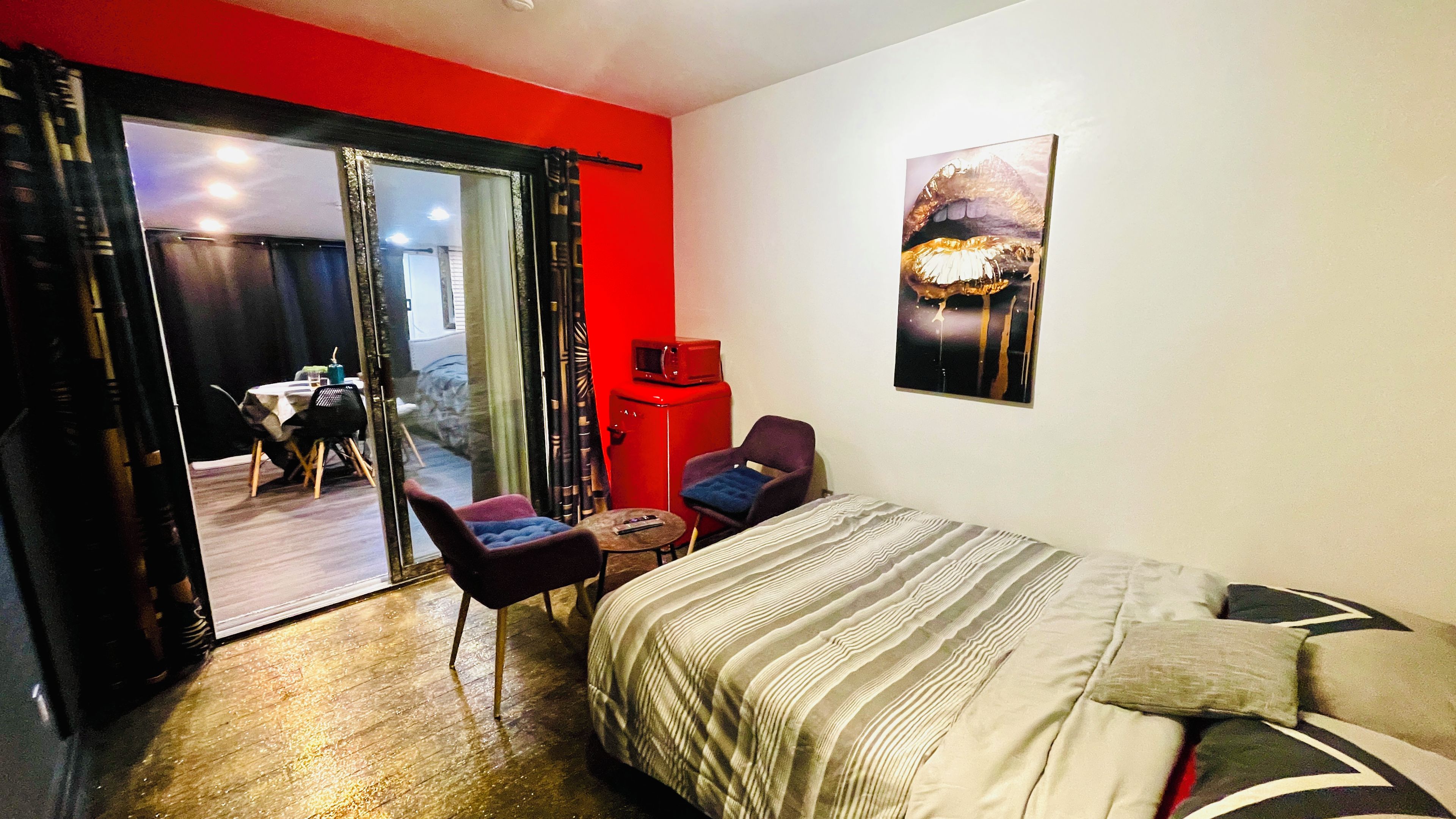A hotel room with a bed, a chair, a small table, a red cabinet, and a sliding glass door leading to a balcony.