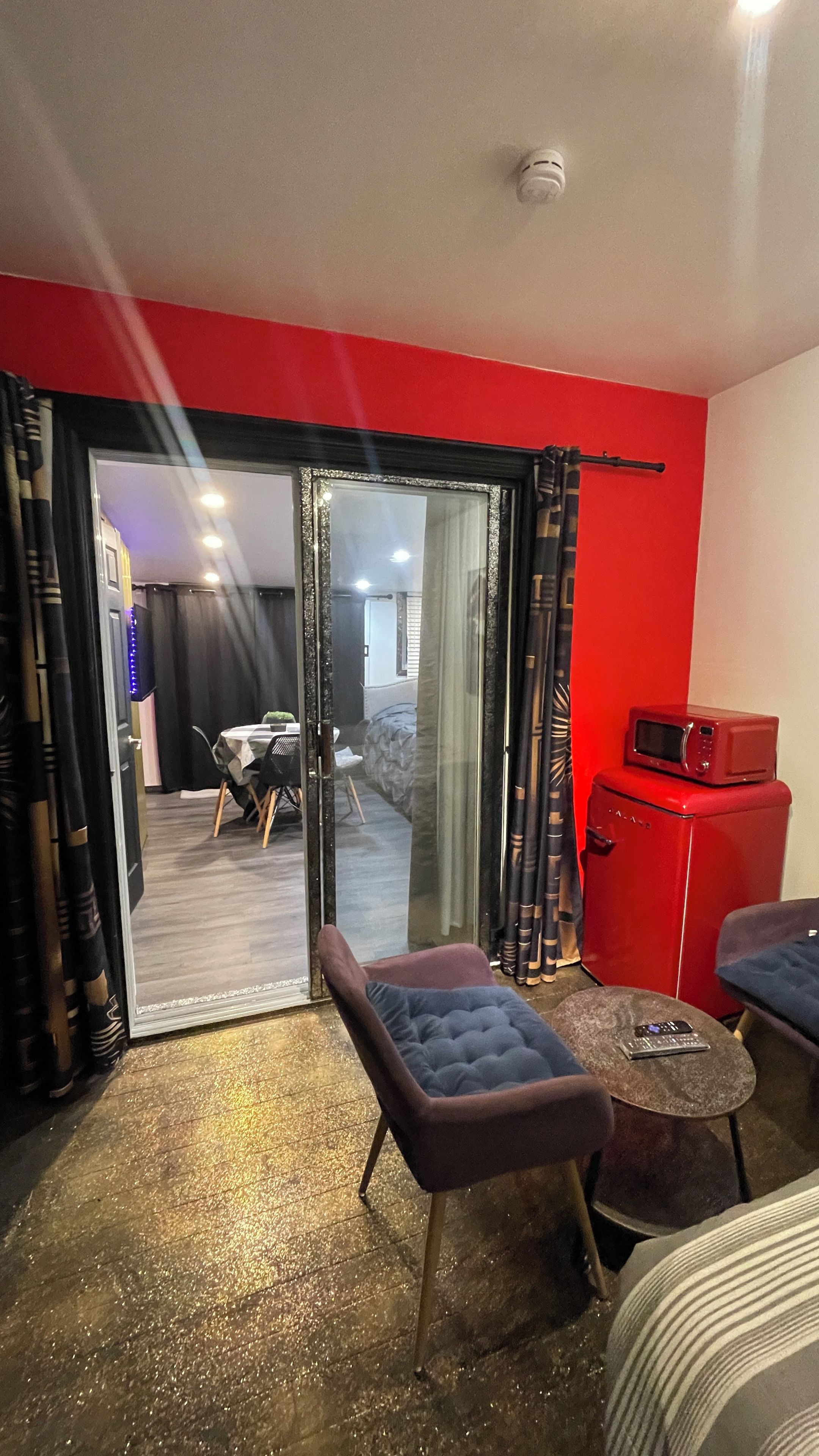 A room with a red wall, a sliding glass door leading to a dining area, a red microwave, and a small table with a remote control.