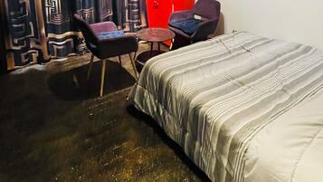 A hotel room with a bed, two chairs, a red wall, and a painting.