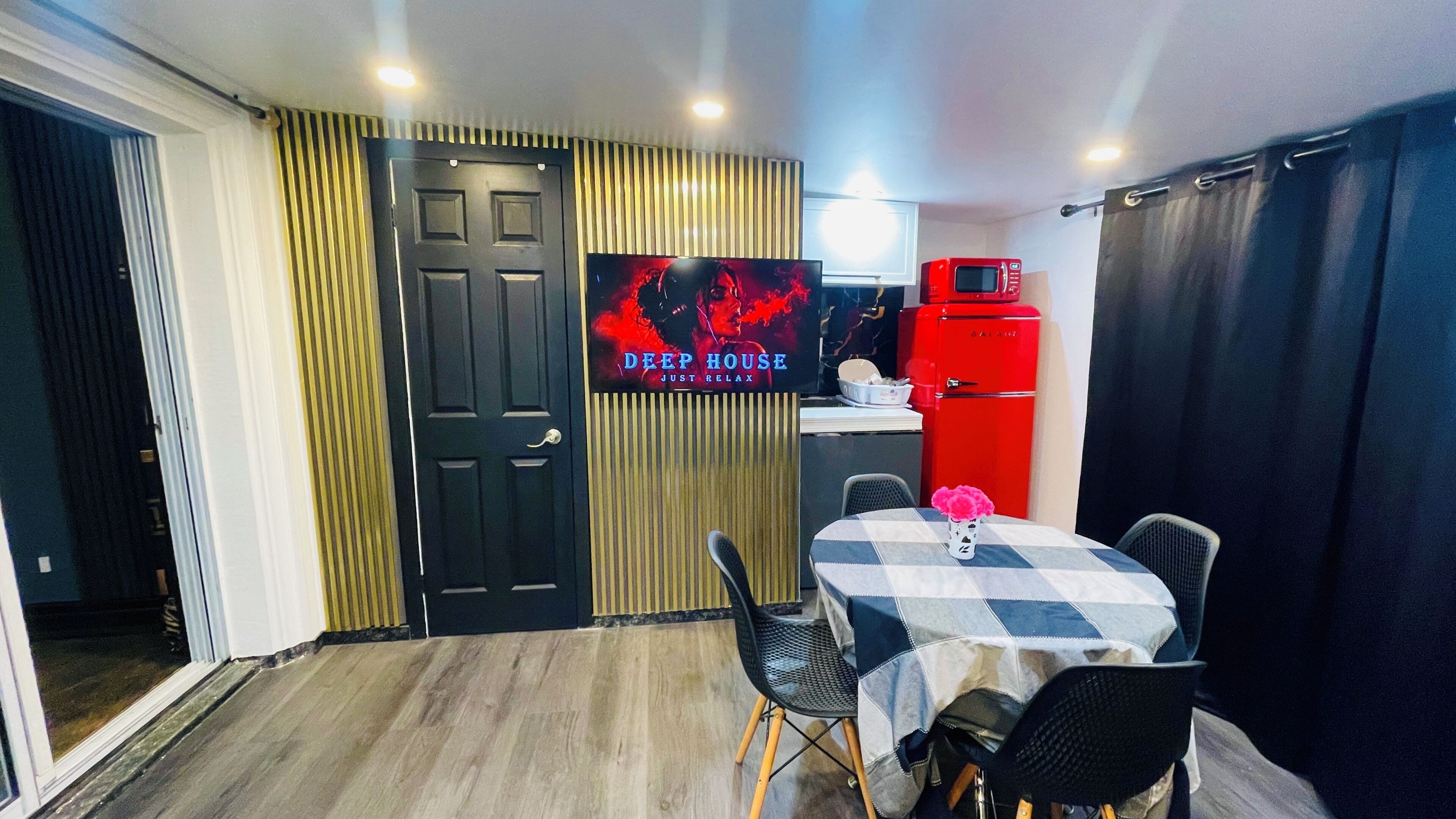 A room with a black door, a television displaying 'DEEP HOUSE JUST RELAX', a red vintage refrigerator, and a table with chairs.