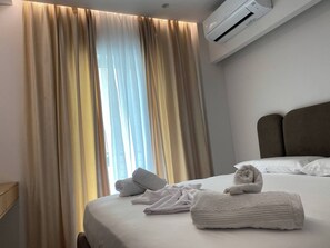 A hotel room with a bed, a window with curtains, an air conditioning unit, and neatly folded towels. - Exquisite Blue Breeze - Free Parking Apartment (Sarandë)