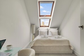 Un dormitorio con tragaluz, una cama con varias almohadas, una mesita de noche con una taza de café y un ordenador portátil.