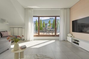 Ein modernes Wohnzimmer mit einem großen Fernseher, einer Couch, einem Couchtisch mit Büchern und Blumen sowie einem Balkon mit Gartenmöbeln und Bäumen.