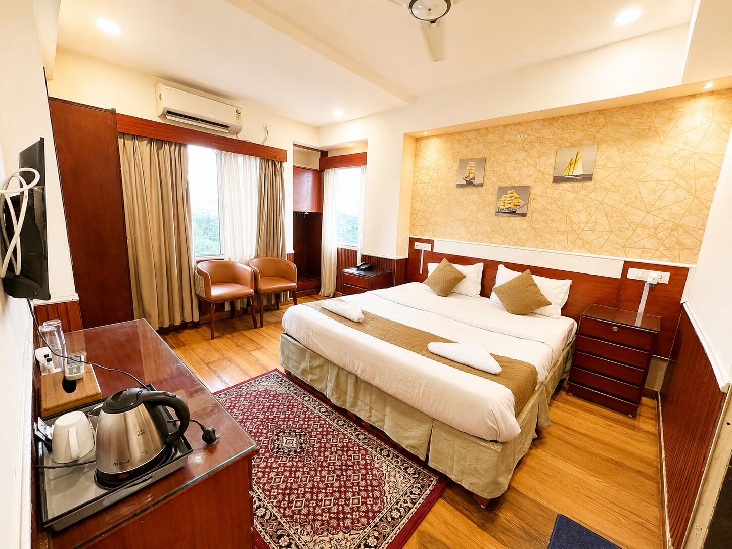 Habitación de hotel con cama, mesitas de noche, escritorio con hervidor, sillas, aire acondicionado y ventana con cortinas.