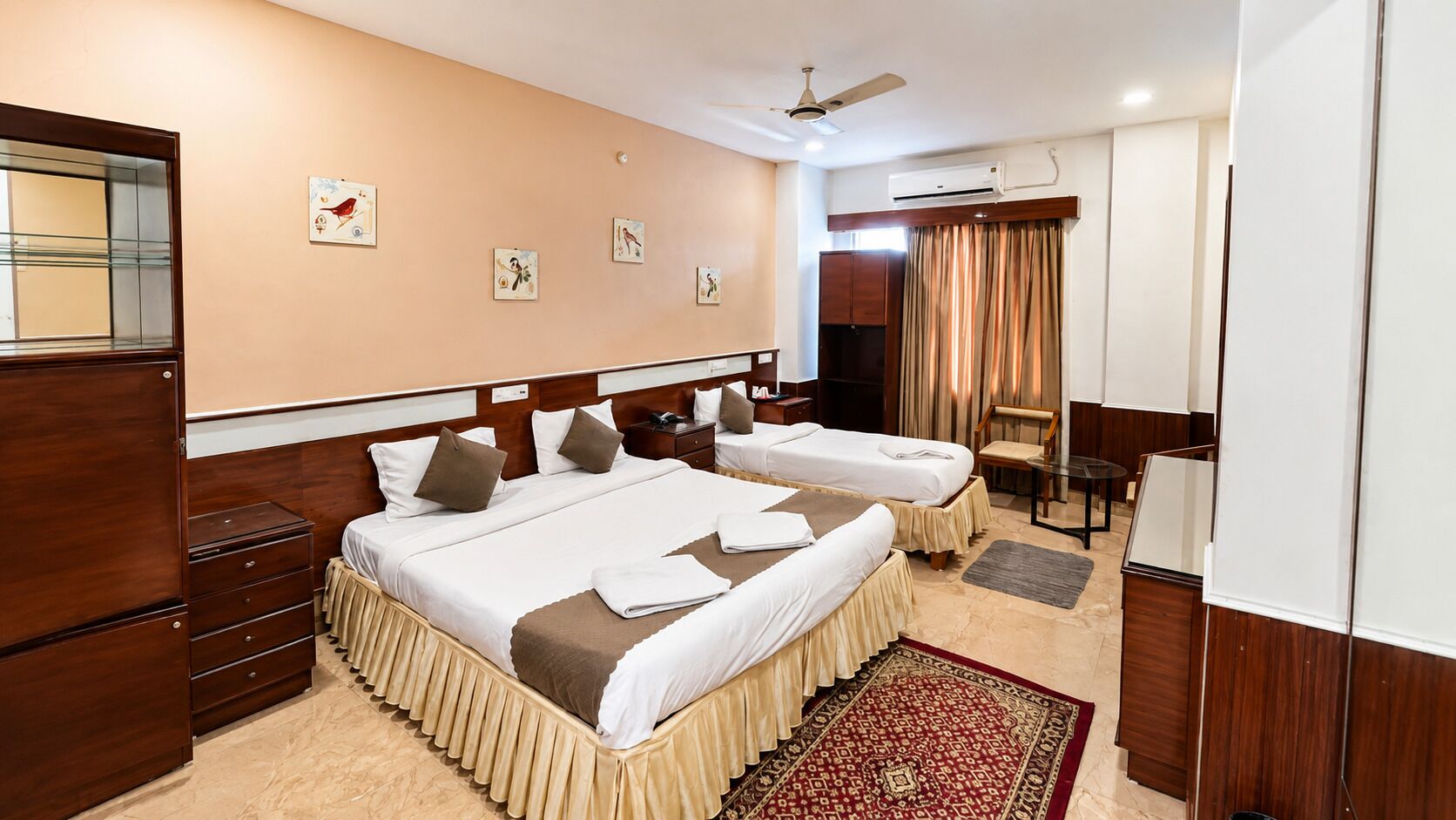 A hotel room with two beds, a desk, a chair, a wardrobe, and an air conditioning unit.