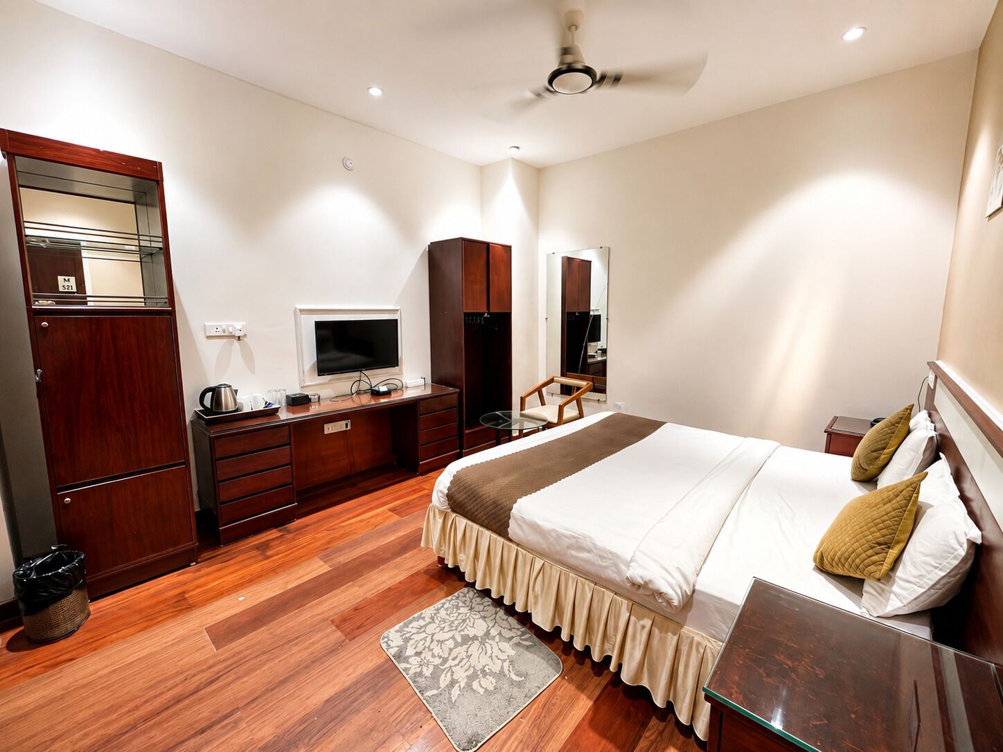 A hotel room with a bed, a desk with a television, a wardrobe, and a ceiling fan.