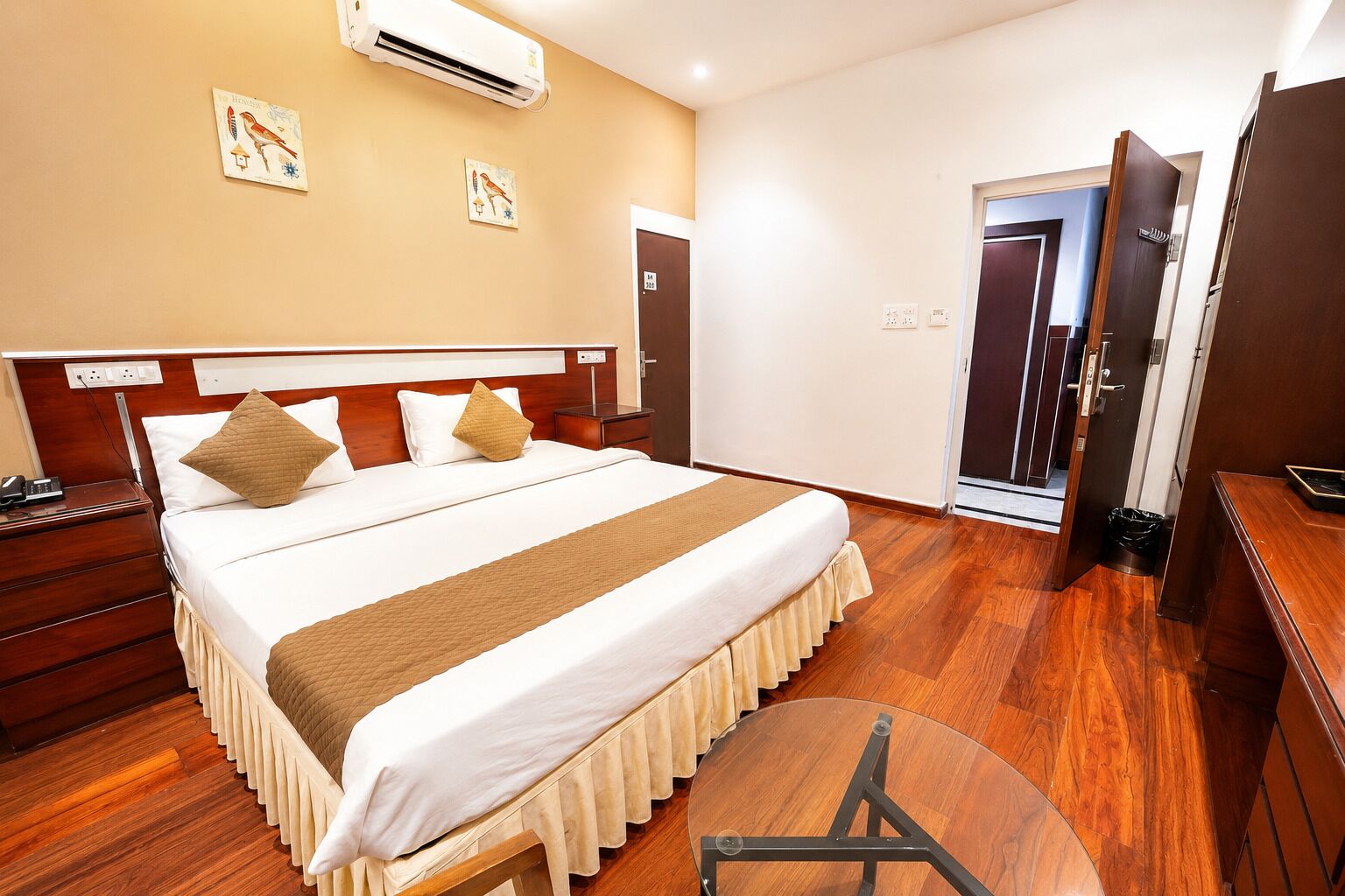 A hotel room with a large bed, bedside tables, a round table, and an air conditioning unit.