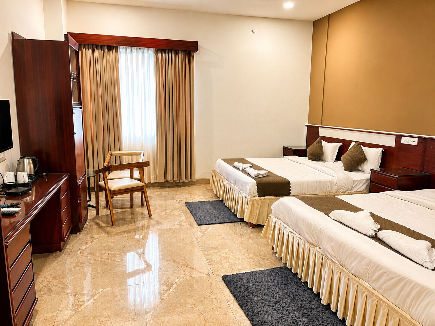 A hotel room with two beds, a desk with a chair, a television, and a large window with curtains.
