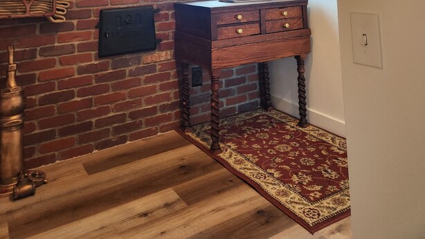 Een kamer met een bakstenen muur, een houten bureau met lades, een raam en een kamerkleed met dessin.