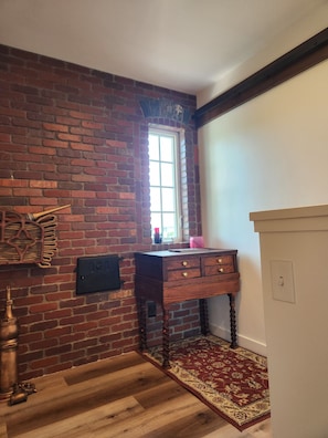 A room with a brick wall, a wooden desk with drawers, a window, and a patterned rug.