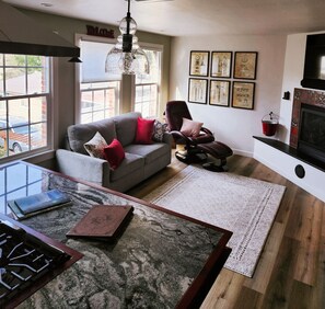 A modern living room with a glass coffee table, a grey sofa with red cushions, a dark leather chair, and a fireplace.