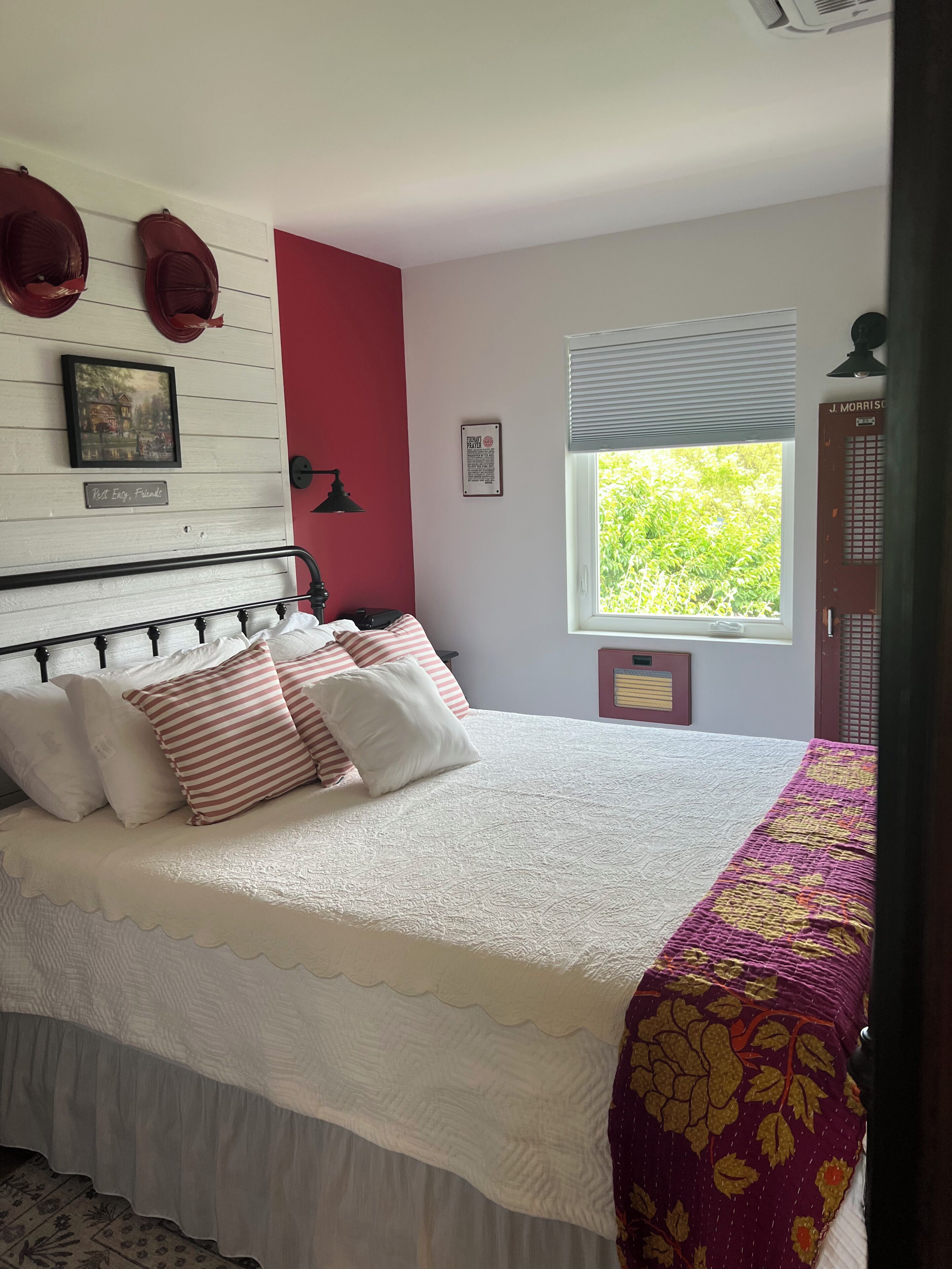 A bedroom with a bed, pillows, a window with a view, a wall-mounted lamp, and a framed picture.
