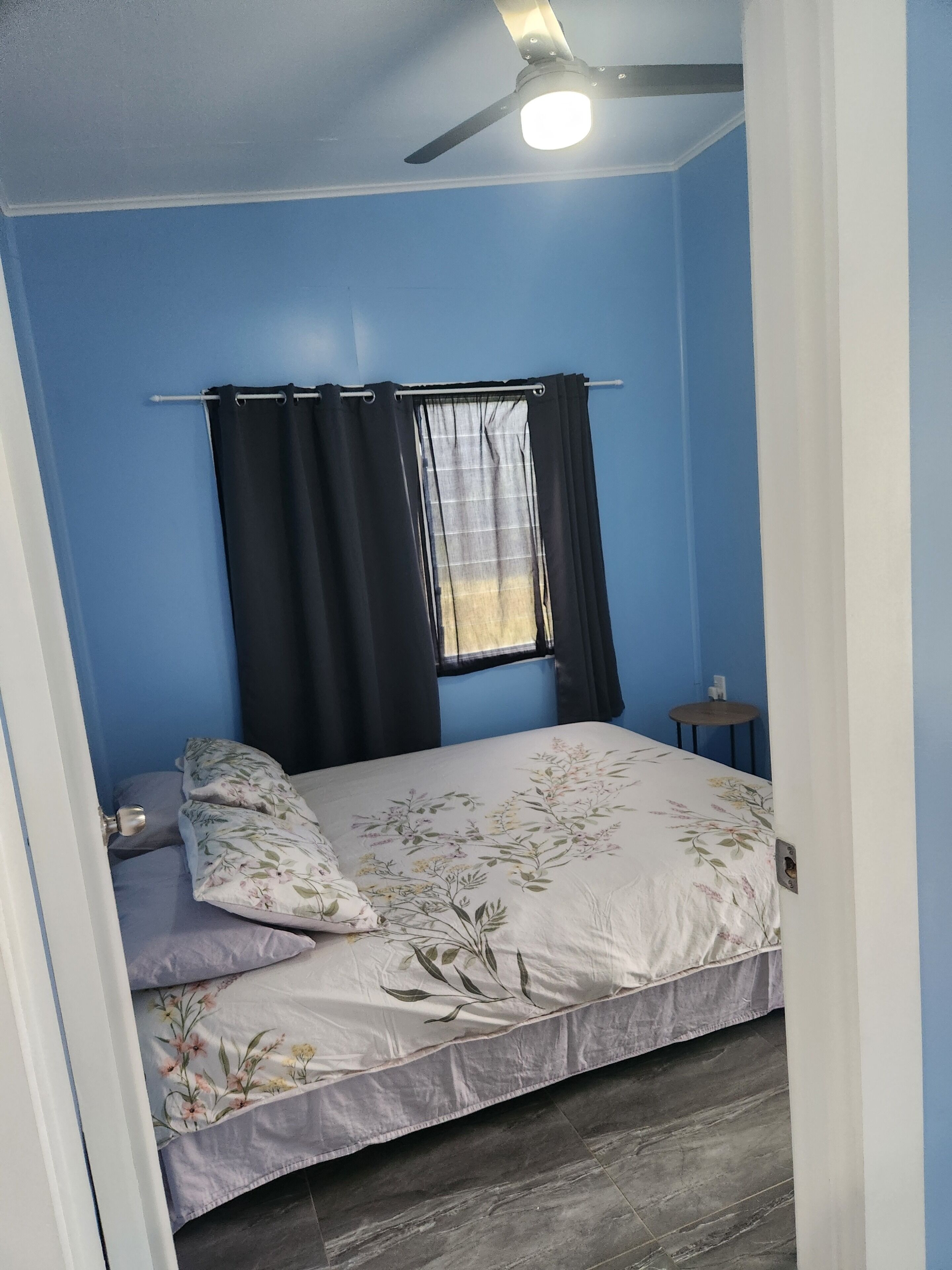 A bedroom with a bed, a window with curtains, a ceiling fan, and a small table.