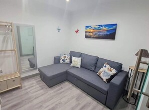 A modern living room with a grey sectional sofa, a glass door leading to a balcony, and a painting of a sunset on the wall.