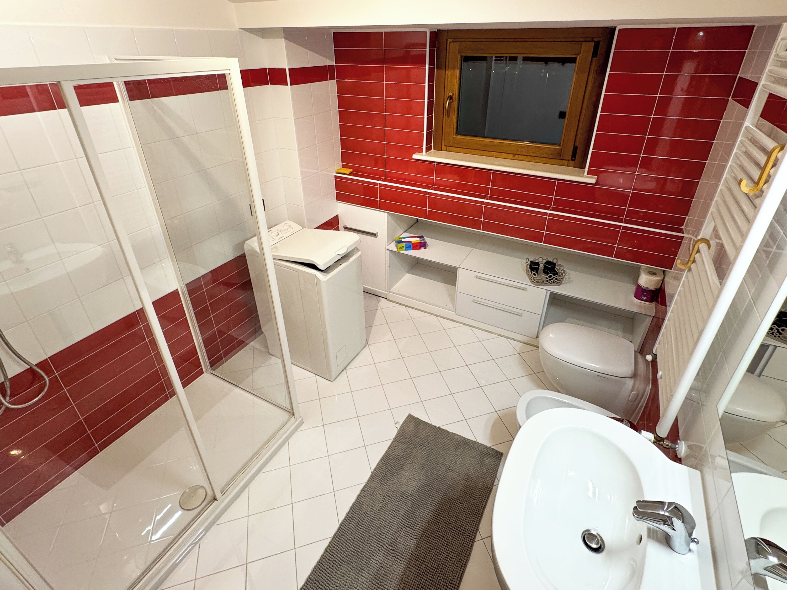 A bathroom with a glass shower enclosure, a white sink, a toilet, and a red-tiled wall.