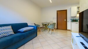 Ein modernes Wohnzimmer mit einem blauen Sofa, einem Esstisch mit Stühlen, einer Küchenzeile mit Mikrowelle und einer Tür zu einem weiteren Raum.