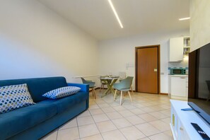 Ein modernes Wohnzimmer mit einem blauen Sofa, einem Esstisch mit Stühlen, einer Küchenzeile mit Mikrowelle und einer Tür zu einem weiteren Raum.