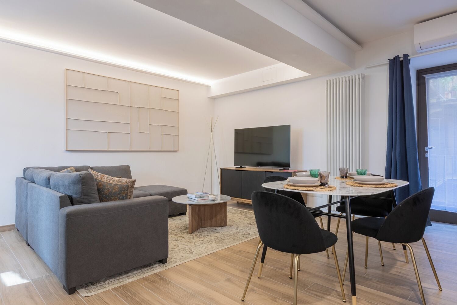 Una sala moderna con un sofá gris, una mesa de centro, una mesa de comedor con sillas, un televisor de pantalla plana y una gran obra de arte abstracto en la pared.