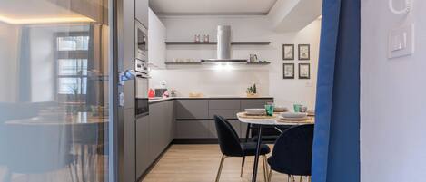 A modern kitchen with a dining area, featuring a glass partition, stainless steel appliances, and a small dining table with chairs.