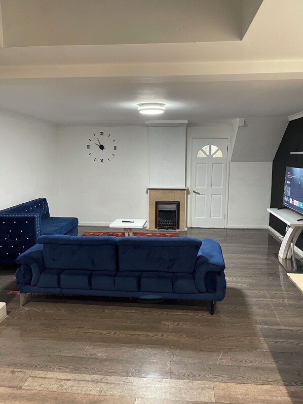 A living room with a blue sofa, a fireplace, a clock on the wall, and a television. - Elites Apartment – Spacious 4-Bedroom Home with Free Parking (London)