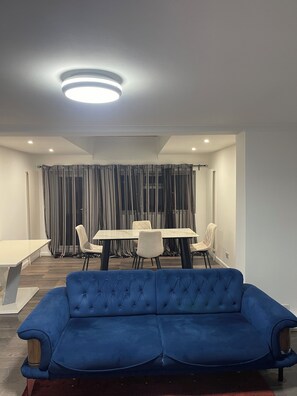 A modern living room with a blue sofa, a dining table with chairs, and a ceiling light fixture. - Elites Apartment – Spacious 4-Bedroom Home with Free Parking (London)