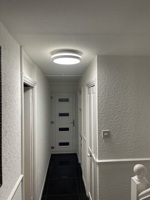 A narrow hallway with a white door at the end, a light fixture on the ceiling, and a textured wall on the right. - Elites Apartment – Spacious 4-Bedroom Home with Free Parking (London)