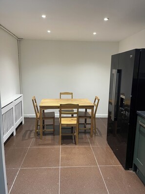 A dining area with a wooden table and chairs, a black refrigerator, and a white radiator. - Elites Apartment – Spacious 4-Bedroom Home with Free Parking (London)