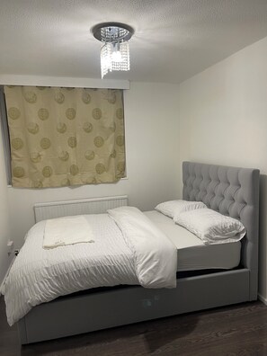 A bedroom with a bed, a radiator, a curtain, and a ceiling light fixture. - Elites Apartment – Spacious 4-Bedroom Home with Free Parking (London)