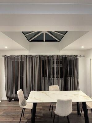 A dining area with a table and chairs, a skylight, and curtains. - Elites Apartment – Spacious 4-Bedroom Home with Free Parking (London)