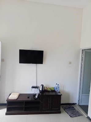 A living room with a flat-screen TV mounted on the wall, a dark wooden entertainment unit with a kettle and remote controls, and a door leading to another room.