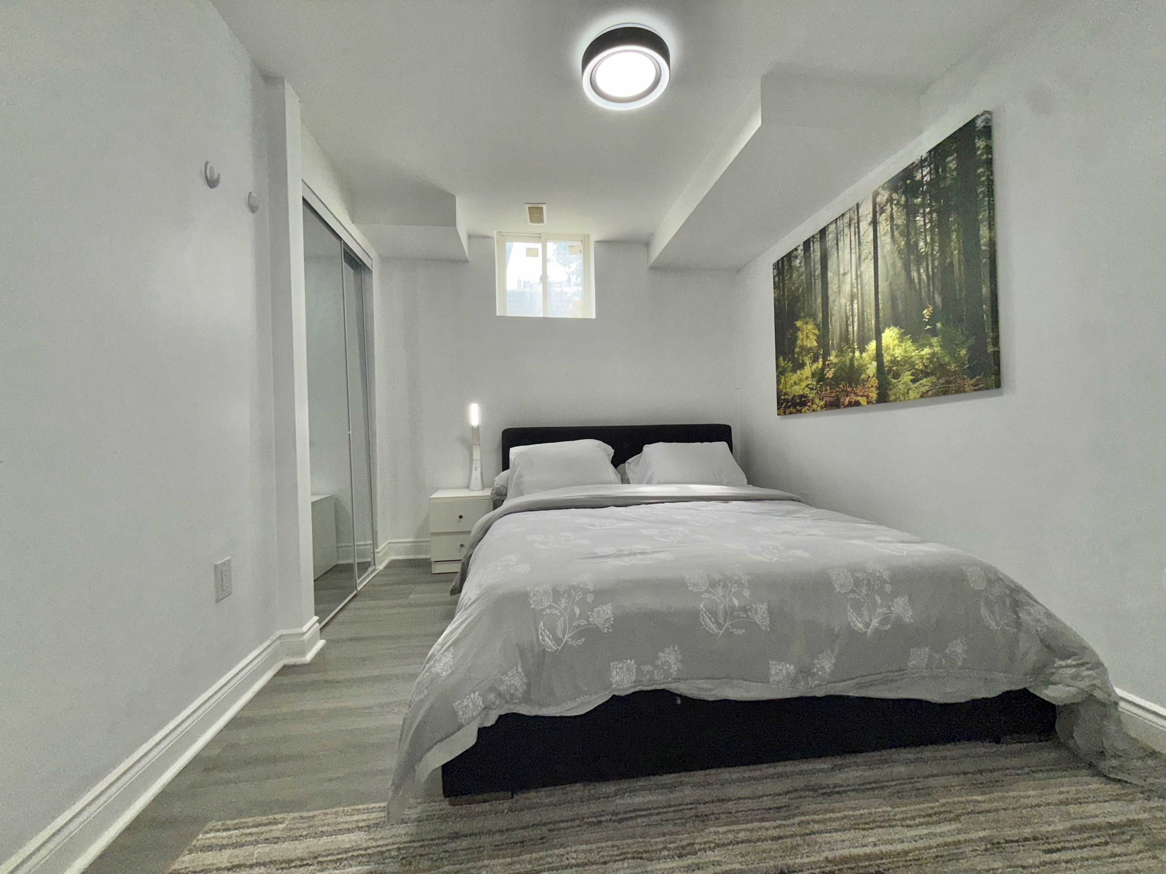 A bedroom with a bed, a nightstand, a painting, and a ceiling light.
