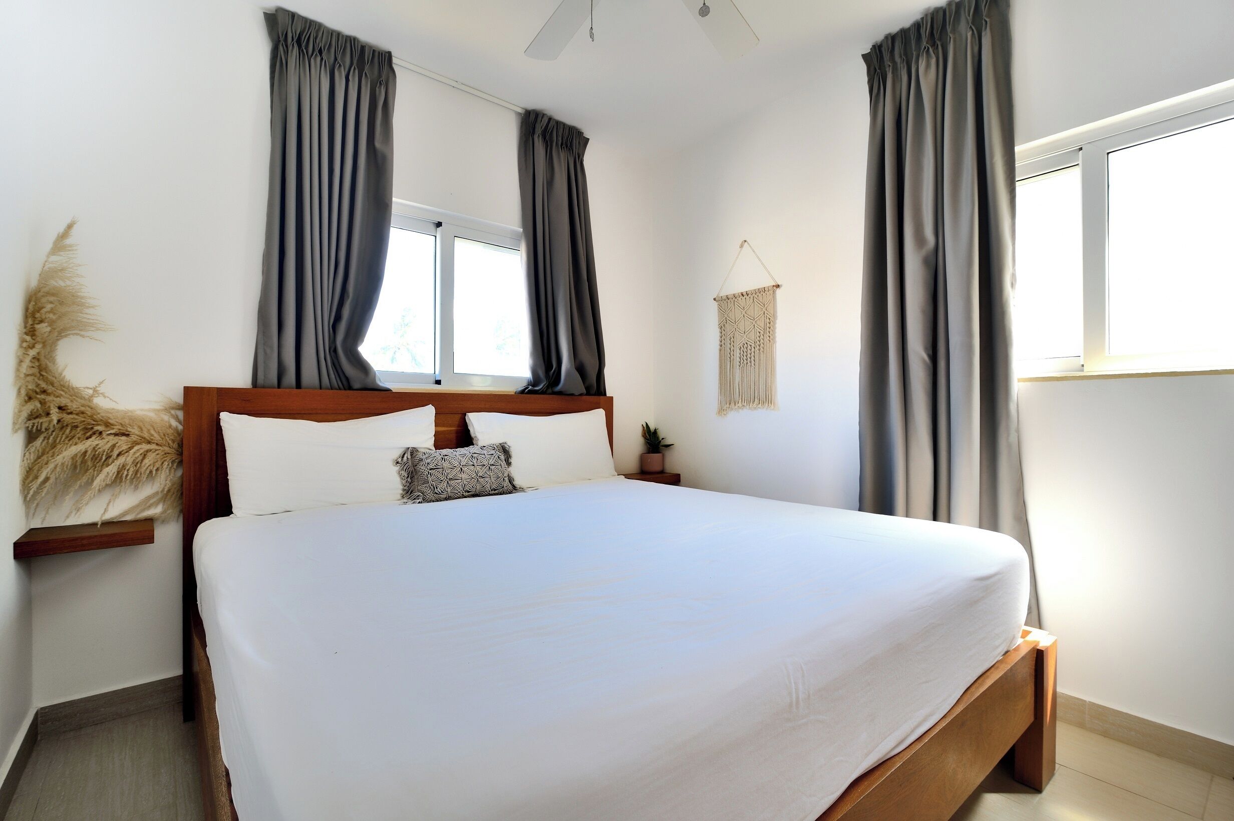 A bedroom with a large bed, two windows with curtains, a ceiling fan, and a wall-mounted macrame piece.