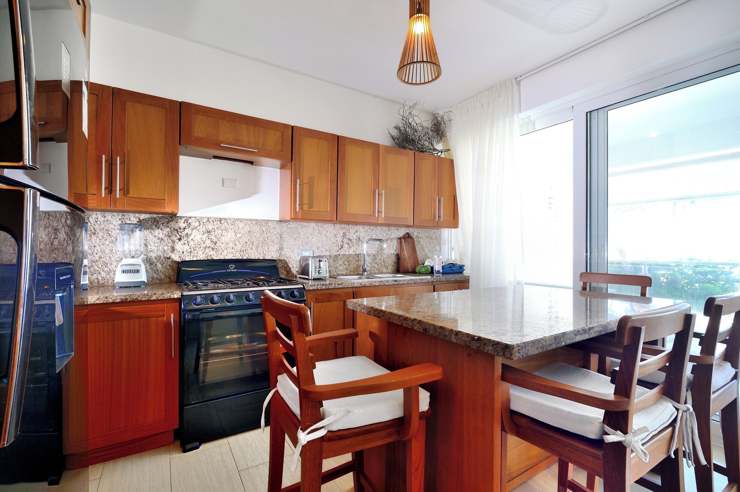 A modern kitchen with wooden cabinets, a gas stove, a built-in oven, and a granite countertop. There is a dining area with wooden chairs and a large window.