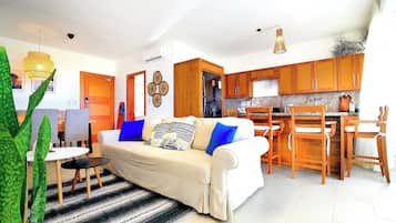A modern living room with a sofa, a dining area with wooden chairs, a kitchen with wooden cabinets, and a large plant.