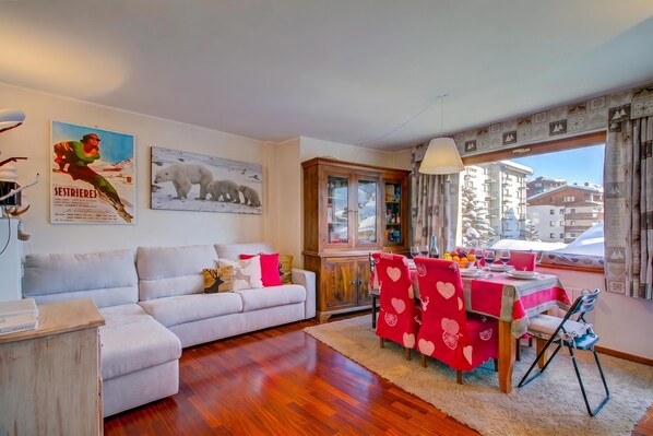 A living room with a sofa, a dining table with chairs, a window with a view of buildings, and a cabinet with glass doors. - Appartamento Venus 11 (Sestriere)