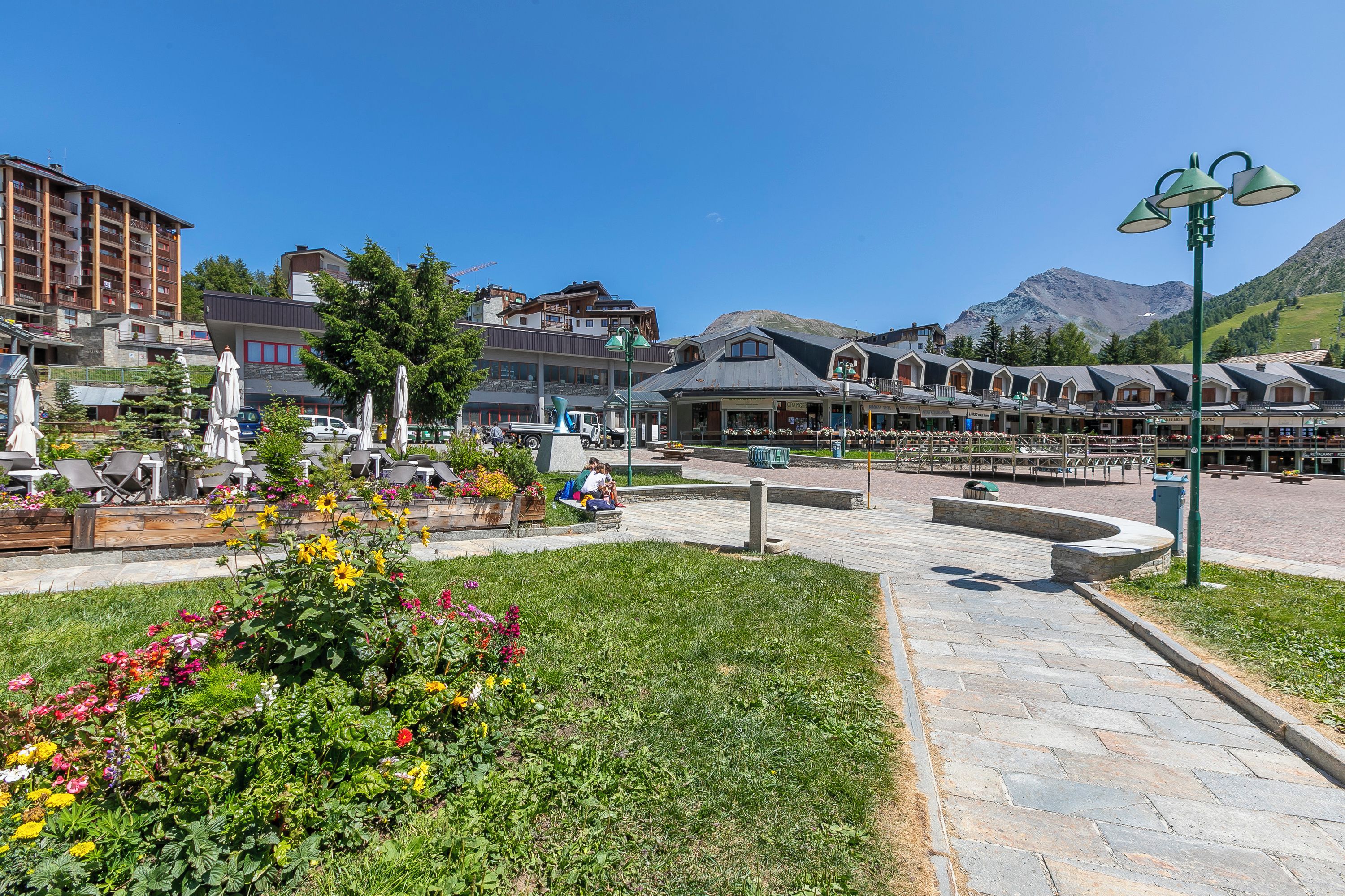 A town square with a central fountain, surrounded by buildings with balconies and a mountain in the background.